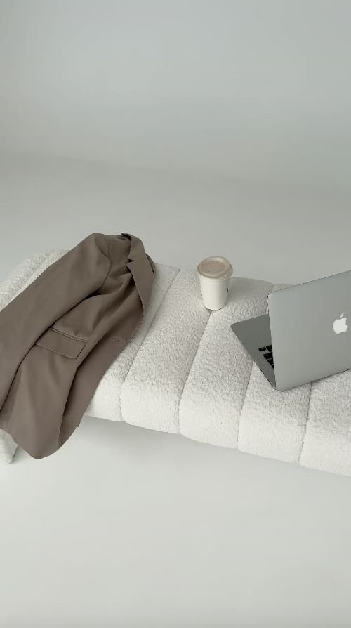 A jacket, coffee cup, and open laptop arranged on a white textured bench in a minimalist office setting